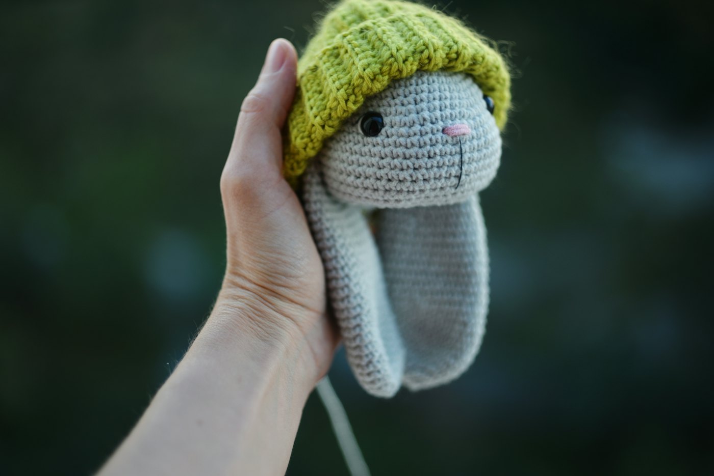 A hand holding a crocheted amigurumi bunny wearing a green hat