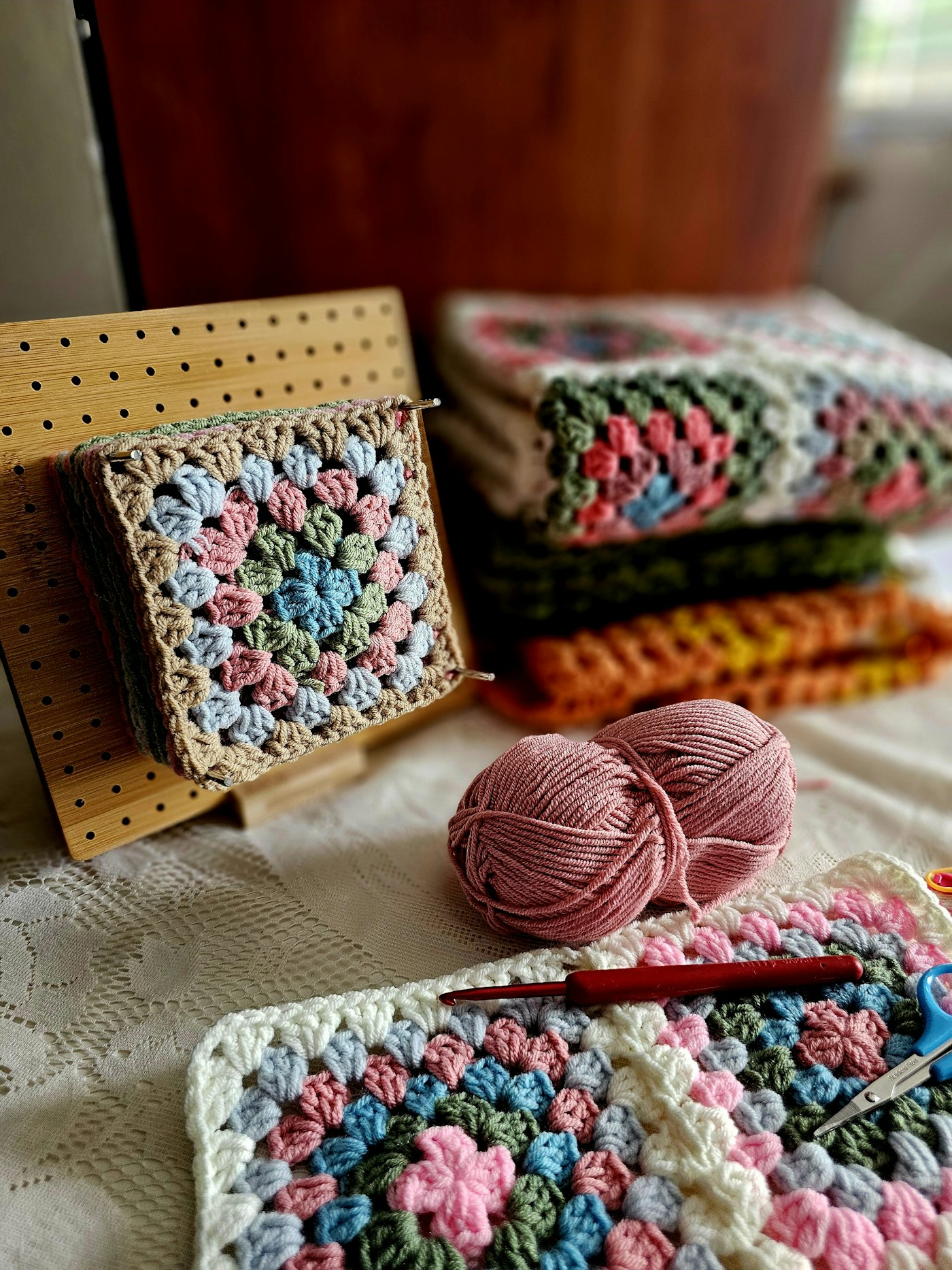 A pastel granny square on a pegboard beside stacked crochet blankets, pink yarn and a crochet hook