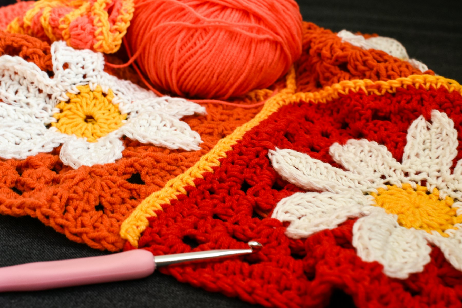 Red and orange crochet squares with white daisy motifs, a ball of orange yarn and a pink crochet hook